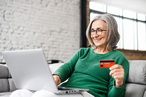 An image of a woman using her laptop. She holds a bank card with her left hand and is wearing a green jumper.