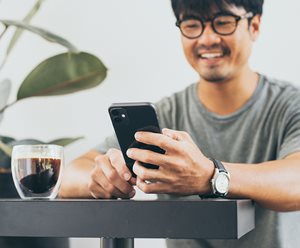 An image of a man sitting at a desk using his phone. The man wears glasses and is smiling with a drink next to his hand.