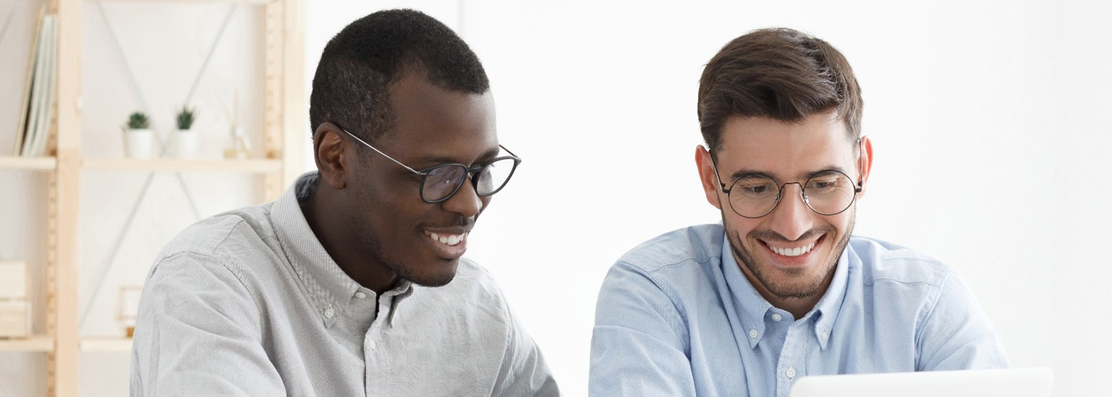 An image of two men sitting a desk looking at a laptop and smiling. The man on the left is wearing a grey shirt and black glasses. The man on the right is also wearing glasses and a blue shirt.