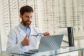 An image of a man in a white coat seated at a desk with a laptop. The man is in an optical practice with many frames displayed on the shelves behind him. The man looks at his laptop screen and has a thumbs up.