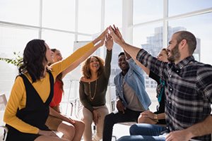 An image of a group of people sitting in a half circle. Each person has their hand raised up to meet each other's hands in the middle.