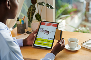 An image of a woman shopping for sunglasses using a tablet. The woman sits at a desk with a cup of coffee next to her.