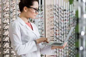 an image of a woman in a white coat standing and holding a laptop. The woman is in an optical practices with many frames displayed on the shelves.