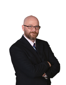 A photo of Dan Baker. He is wearing a pair of black glasses and a white shirt with a blue and purple tie and a black blazer.