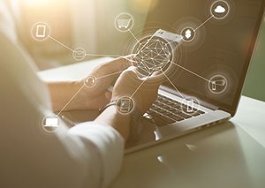 An image of a person using their smartphone in front of a laptop.
There are several icons overlaid on the image, creating a visual network that suggests that the person is connected to the internet. The icons include a shopping cart, a cloud, a phone, a mail envelope, and a headset. The icons are connected to each other by lines, and the lines are also connected to the laptop and the smartphone.