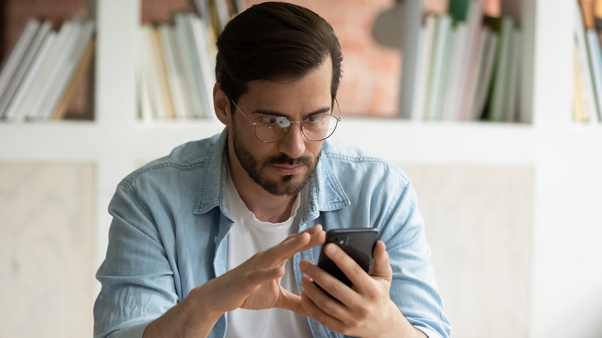 An image of a man using his phone. He is wearing a pair of glasses and a white shirt under a blue long sleeved button up shirt.