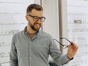 An image of a man wearing a grey shirt and black glasses. He is holding a pair of glasses frames in his hand.