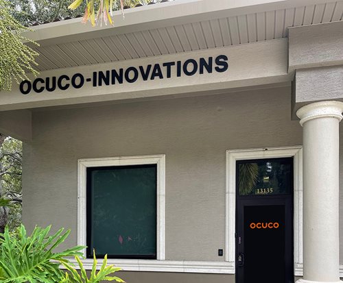 A photo of the entrance to an office building. The walls are beige and above there is a sign that says 'OCUCO-INNOVATIONS' in black. The door to the entrance is black and has 'OCUCO' logo in orange in it.