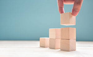 The image shows a hand placing a wooden block on top of a stack of three wooden blocks. A single block is on the left side of the image, a stack of two blocks is to the right of that, and a stack of three blocks is on the far right.