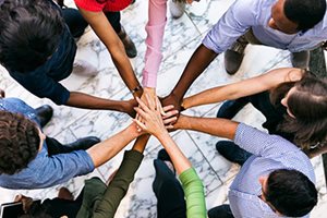 An image of a group of people in a circle with their hands joined together at the center. The people in this image all have different skin tones, displaying diversity.