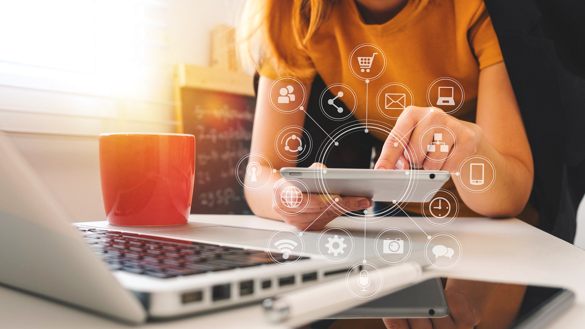 An image of a hand holding a notebook with a laptop on a desk. Overlaid on the image are white icons of cameras, gears, phones, globes, shopping carts and a WiFi connection